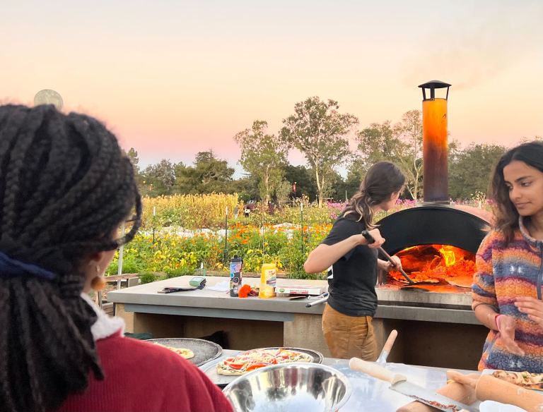 Two students cooking and serving pizza, while a third stands in front of them waiting for the pizza.