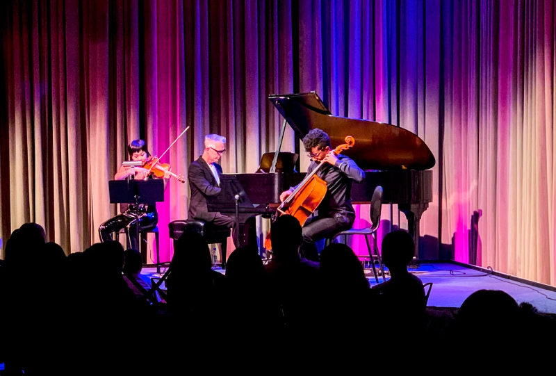 a photo of three musicians performing at a classical concert; left to right, they respectively play a violin, a piano and a cello