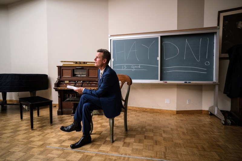 Geoff Nuttall sits in a blue suit on a wooden chair, looking left. He holds a rolled piece of paper and his legs are crossed. Behind him is a blackboard with the words, "HAYDN! ... is good." The word "HAYDN" is very large and underlined. Also behind Nuttall are an antique keyboard instrument and a covered piano.