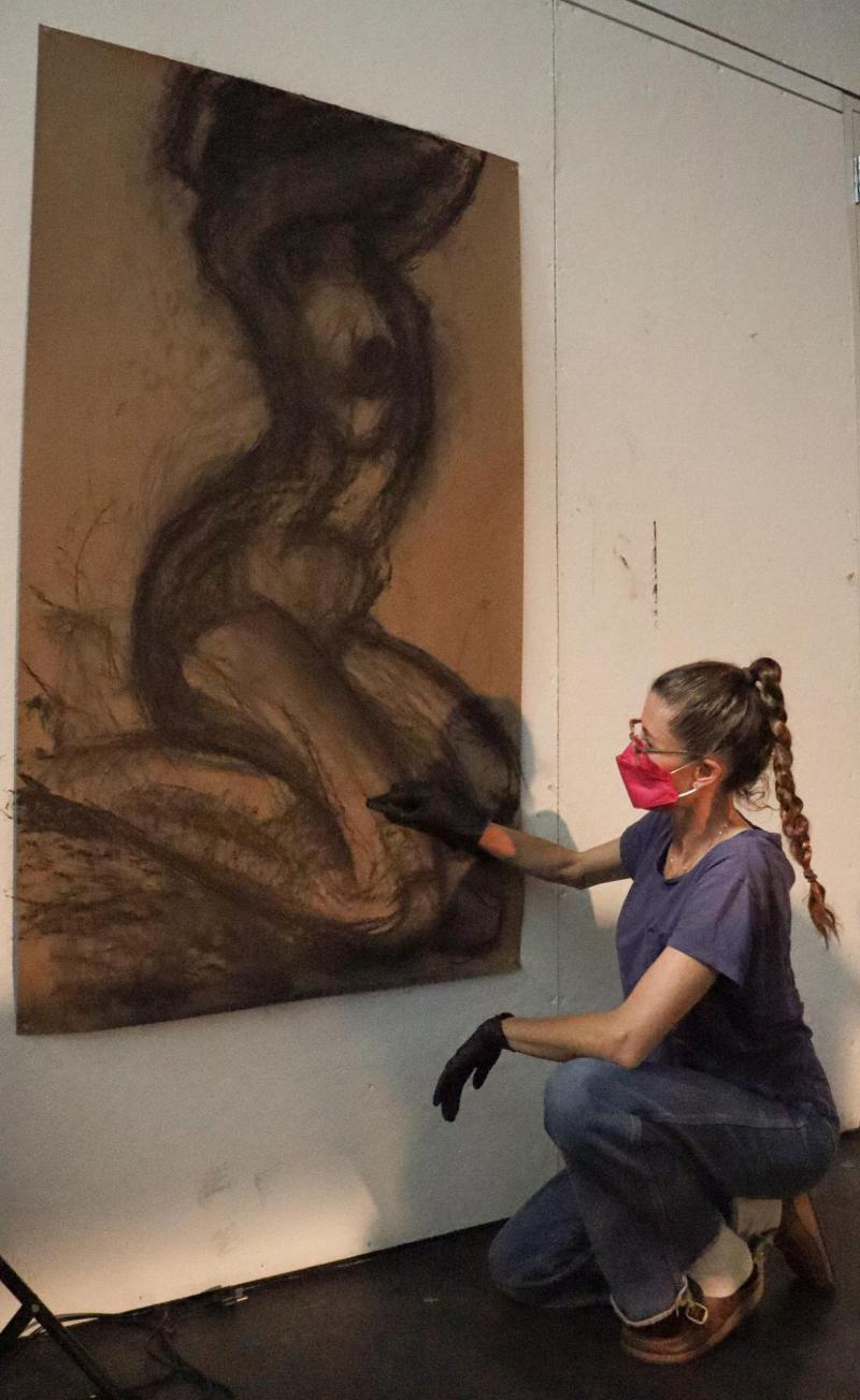 Art professor crouched while drawing on a brown canvas the sketch of a body.