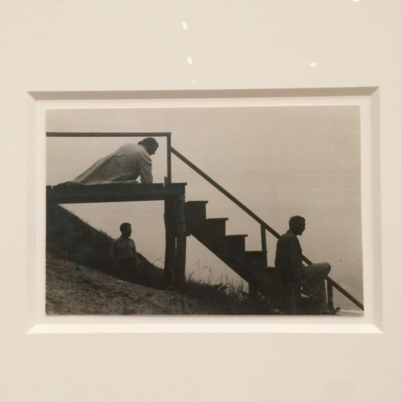 In a monochrome photograph, three figures are positioned around a wooden staircase on a beach. The figures and the staircase are all in profile; one person lies at the top, looking at another sitting on the steps. A third stands under the stairs.