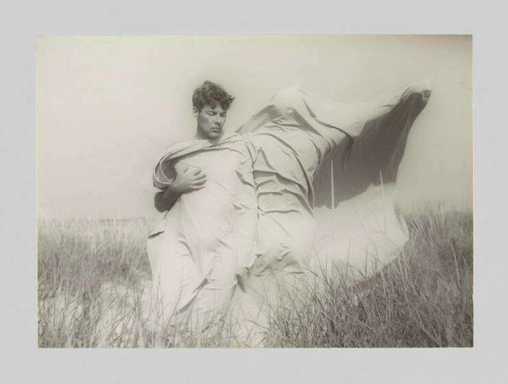 In a monochrome photograph, a man with brushy hair stands with another human figure in a tall grass field. A large white sheet covers the man's torso and the entire body of the other figure, wrinkled and caught by the wind. The figure has an arm stretched toward the sky.