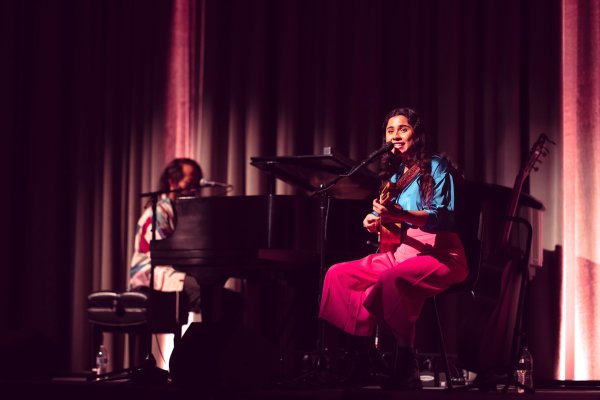 on stage, a person sits at a piano at the left and another sits in front of a microphone holding a guitar
