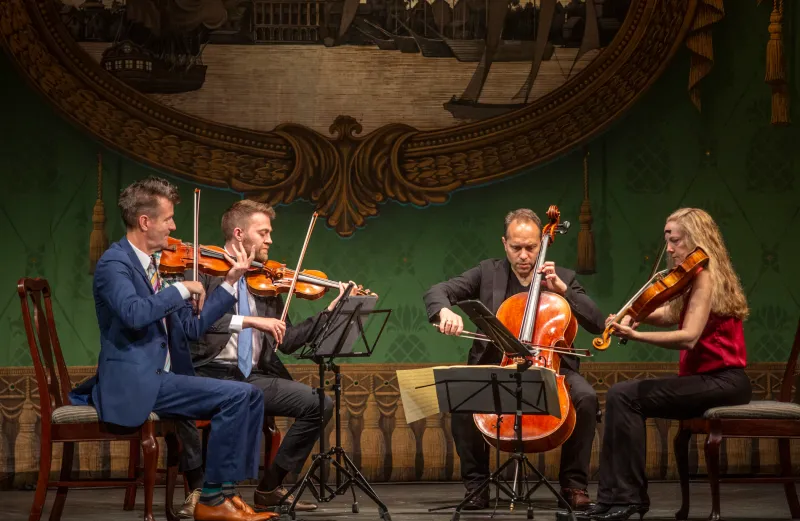 The St. Lawrence String Quartet plays  in a half-circle onstage in front of an ornate green and brown background. The violinists, Geoff Nuttall and Owen Dalby, sit in chairs on the left. The cellist, Christopher Costanza, sits in the middle right. The violist, Lesley Robertson, sits on the far right. Each is playing their instrument actively.