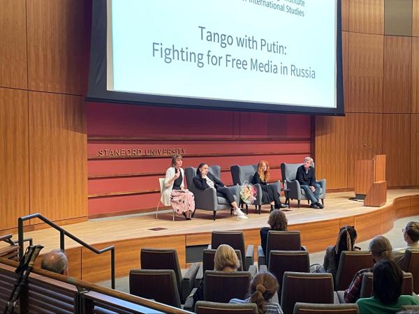 Four panelists, including Sindeeva and filmmaker Vera Krichevskaya, sit on a large wooden stage in blue armchairs. Behind them is a red paneling with the words "Stanford University," and above them is a screen with the projected words: "Tango with Putin: Fighting for Free Media in Russia." Several audience members sit in seats below them.