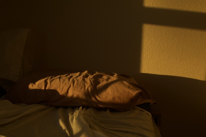 A formless figure lays in bed, undefinable beside its feet. Sunlight peeks in through a window.