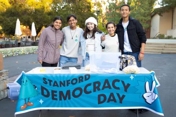 Five students stand in front of a booth with a large blue sign that reads Stanford Democracy Day.