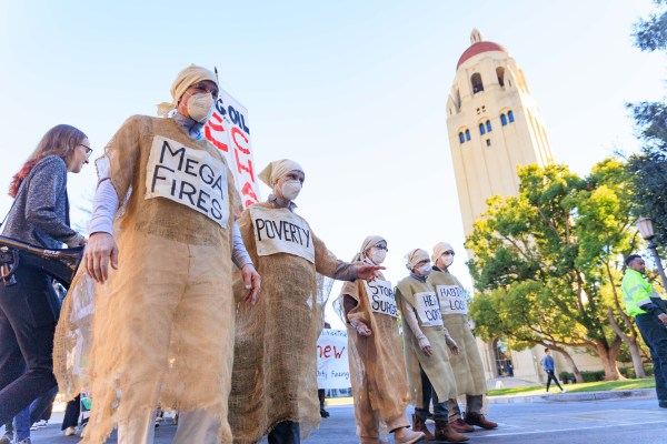 People dressed in burlap smocks bearing signs that read "mega fires," "poverty," ,"storm surge," "habitat loss," and other environmental activism phrases demonstrate outside the Hoover Institution with a tall Hoover Tower in the background.