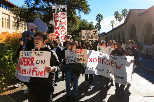 Students carry signs reading phrases like "shut the Doerr on fossil fuel" and "stop fossil fuel capture" while marching beside Main Quad at Stanford University.