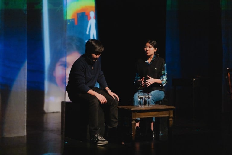 On a darkened stage, a man dressed in a vest and jeans questions another. The other bows his head down in contemplation.