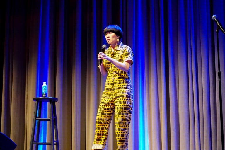 A woman in yellow stripped and dotted overalls holds the mic in her hands, stands on Bing stage, and stares at the audience with a surprised look