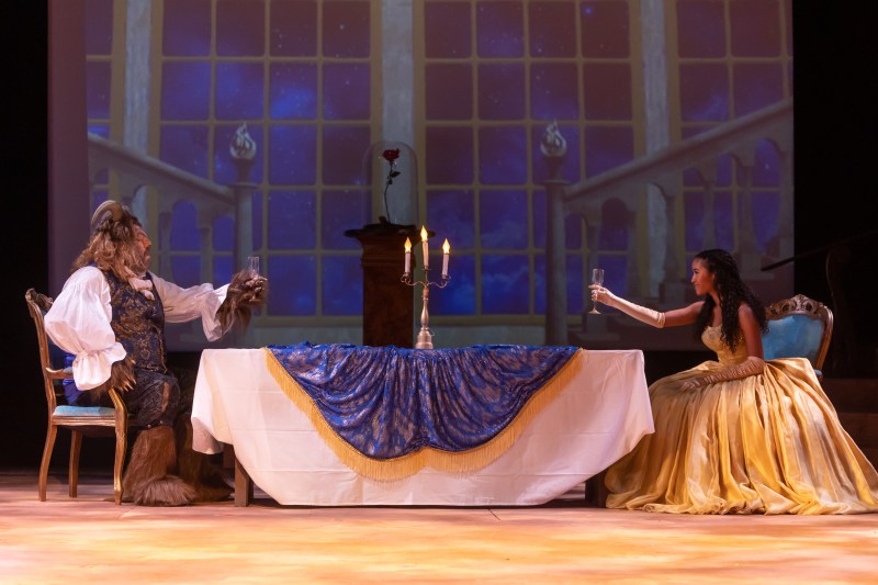 The beast and Belle raise the glass to each other at a large table with dark blue table cloth and archaic candles on top.