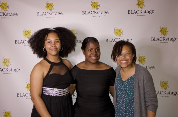 Three women stand in front of a white screen with prints of "BLACKstage"