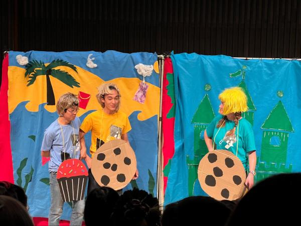 Against a colorful backdrop, a man wearing a cardboard muffin holds the hands of another with a cookie. A woman wearing a yellow wig and a cookie cardboard stares at them as if in anger