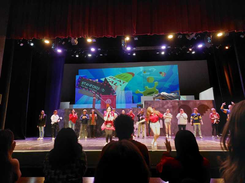 Actors wearing red stand on stage. The background of the stage is a screen displaying Hoover Tower and “BEAT CAL”