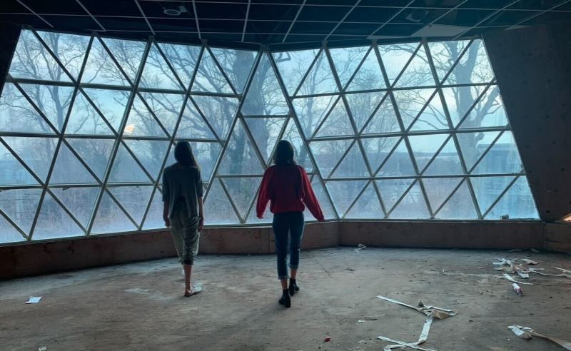 Two women stand inside a geodesic dome. Outside, through the windows, a forest is visible.