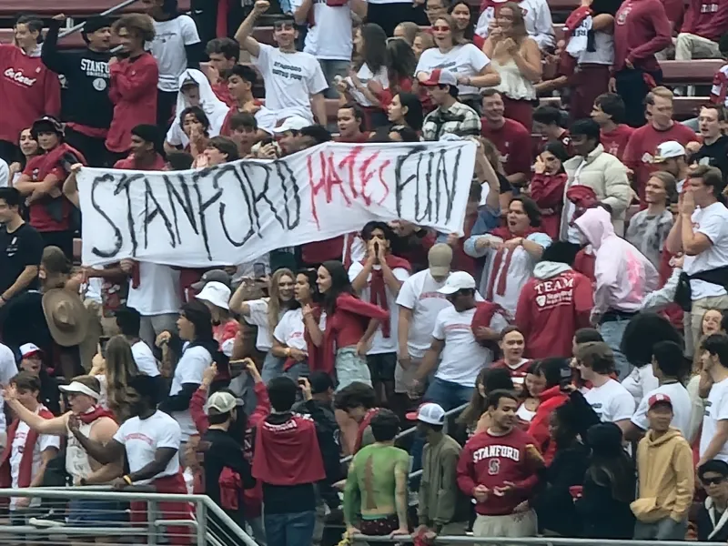 A group of Stanford students standing in the stands of the Stanford Football stadium hold up a large, white banner that reads "Stanford Hates Fun" in black and red font.