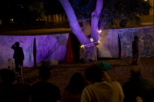 A crowd of people watch an outdoor production of Waiting for Godot. There are two actors standing on the left and right and in the center there is a tree lit up by purple lighting. On the tree there is a sign reading, "Next Day. Same Time. Same Place."