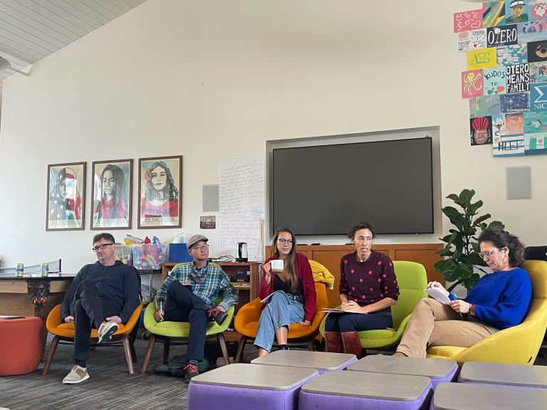 Five people in casual clothing, men and women, sit on colorful armchairs in the Otero lounge. Behind them are a TV and posters.