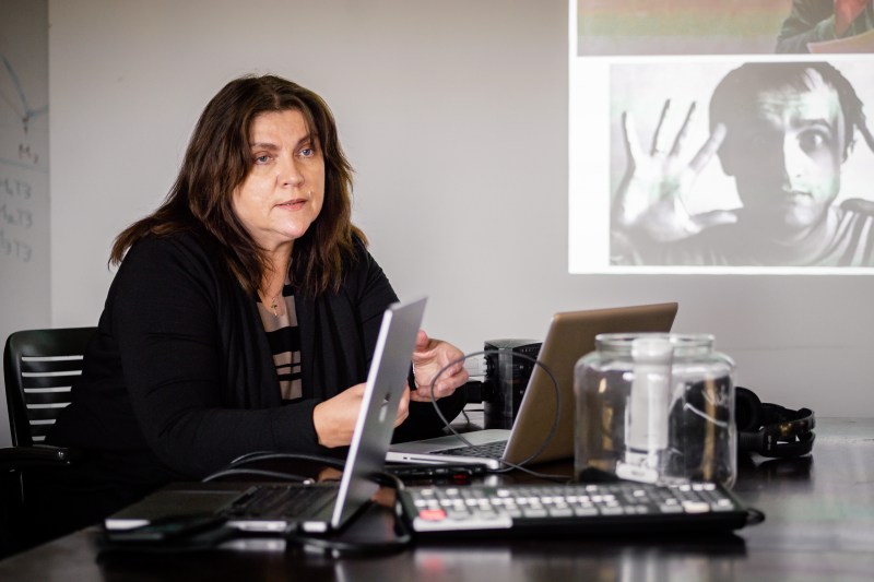 a white person with long brown hair sits at a desk in front of an computer; a presentation slide with a collage of photos is projected on the wall behind her