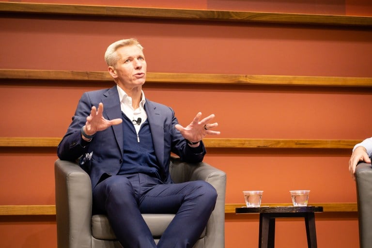 Harry Brekelmans sits in a grey chair on stage, water to his left, with an orange background.