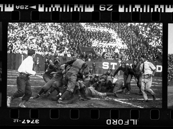 A black and white photo of football players clamoring over each other in a stadium. The photo is surrounded by a frame as if it was part of a film strip.