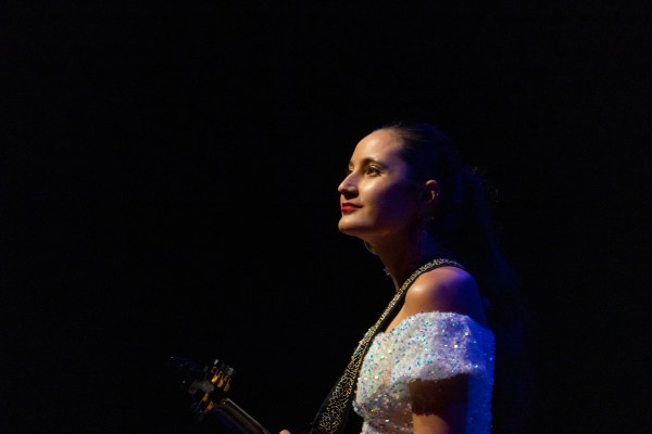 a shoulders-up photo of a woman on a dark stage wearing a white off-the-shoulder dress and looking up toward white lights