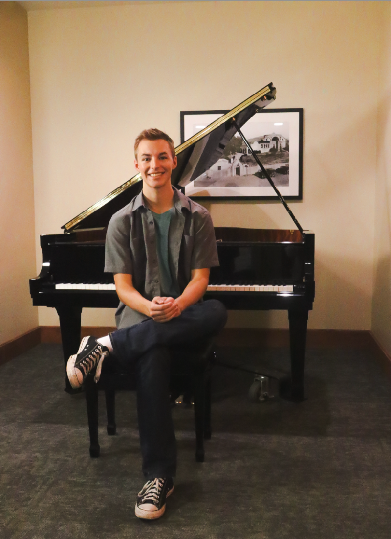 a caucasian person with short hair sits at a piano facing the camera. he wears a dark gray tee shirt and black pants