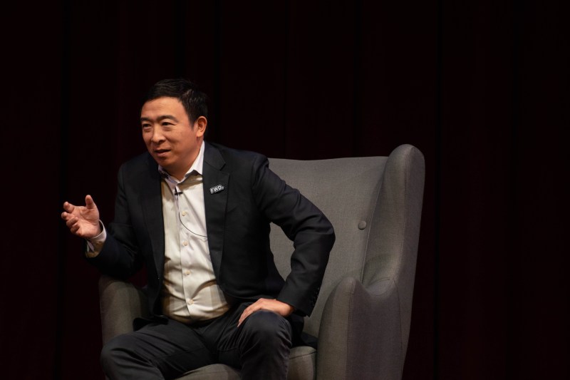Andrew Yang sits in a chair in the middle of the stage at Dinkelspiel Auditorium with his arm extended while speaking to the crowd.
