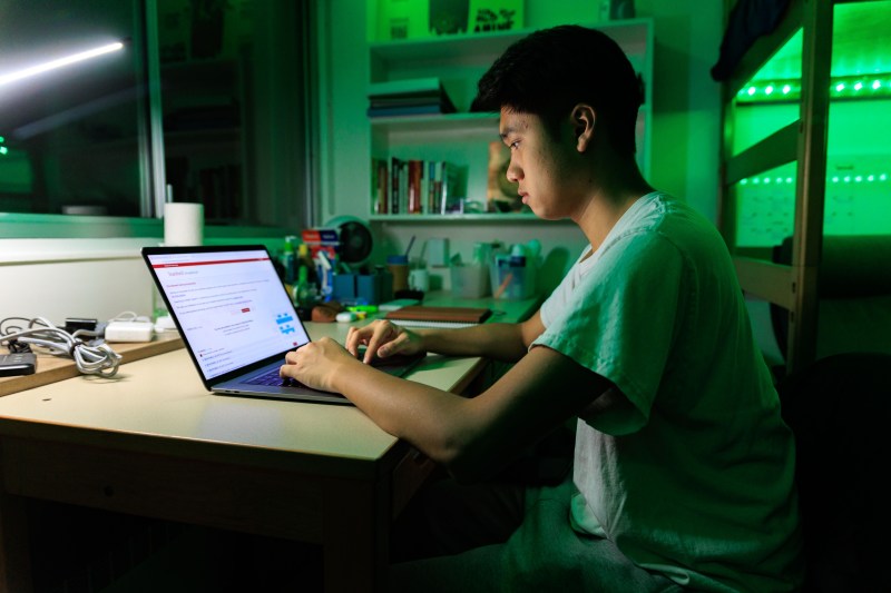 Scott Vu sits at his desk, facing towards the left of the frame. His hands are on the keyboard of his MacBook, which displays the screen of the new SimpleEnroll system. A desk lamp lights Scott's face while green LED strip lights illuminate the background.