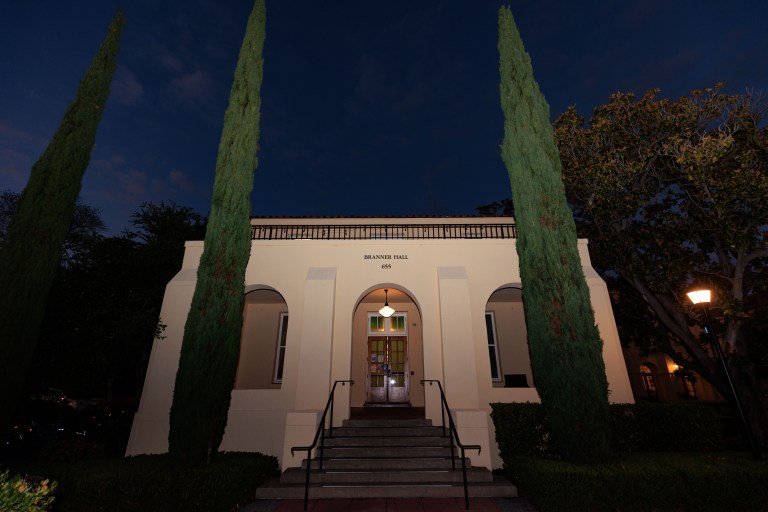 Branner Hall, a dorm, is lit. A deep blue sky above. Three tall, skinny trees out front, with one slightly bushier tree to the right. Stairs lead up to the door, which sits under an illuminated exterior light.