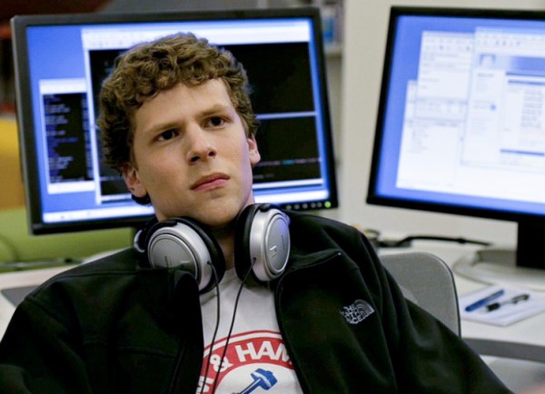 Curly-haired Jesse Eisenberg wears a white t-shirt with a red Arm & Hammer logo, with a black jacket over and headphones around his neck. He sits at a desk with two monitors, facing away from the screens.