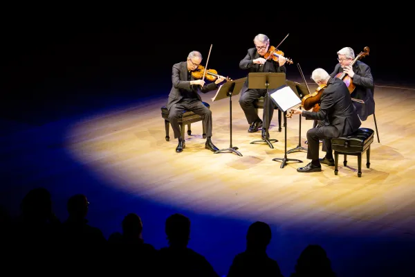 four older people in suits are sit in a semicircle, playing varied string instruments and reading off music stands