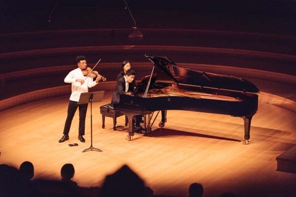 on a lit stage, a young person with short hair plays the violin while standing in front of a black stand. to the right, a young person plays the piano with an intensely focused facial expression.