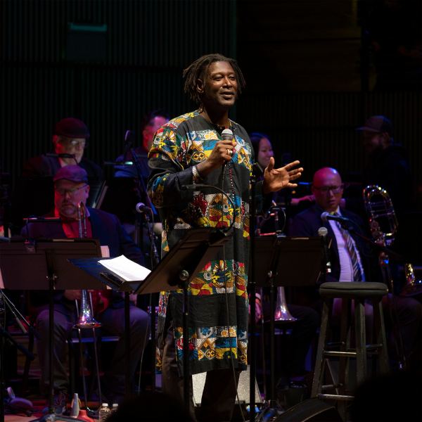 A Black person speaks and gestures before an audience on a dimly lit stage, wearing a long garment with thick black and multicolored stripes