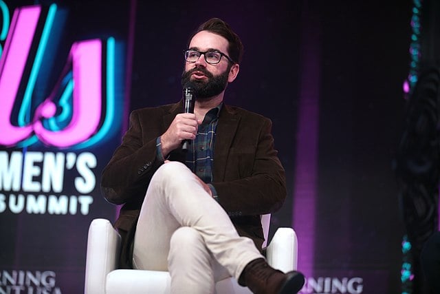 Matt Walsh, a white man with a brown beard and a brown suit, sits and speaks into a microphone on a colorful background