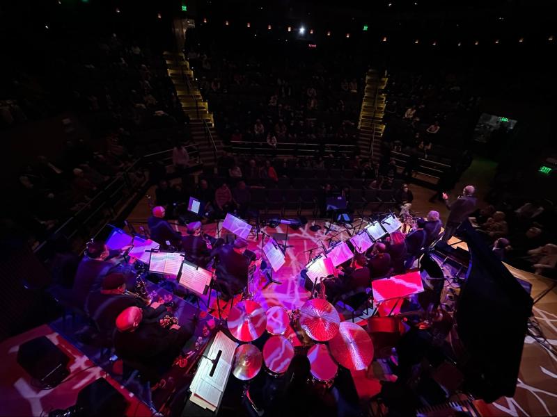 A birds-eye photo of a stage awash in magenta lights; several musicians sit inside two rows music stands; a drumset occupies the middle of the stage