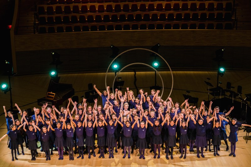 On a brightly lit Bing stage, DiDonato stands with around 50 school-age girls in purple uniforms. The singers have their hands up in celebration.