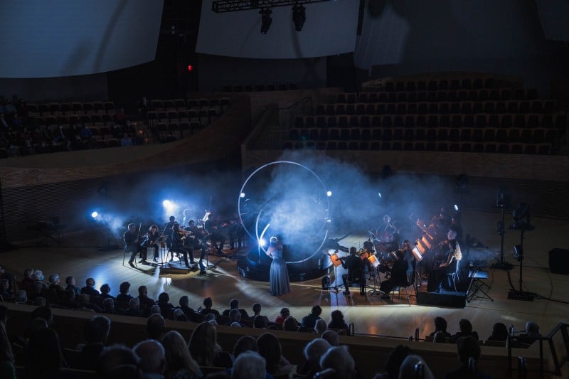 On the stage of the dark Bing Concert Hall, an orchestra surrounds a large black platform with two large upright silver rings. Through haze and bright lights, DiDonato stands in a blue dress at the center.