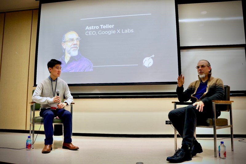 Google X Ceo Astro Teller is sitting down next to a student presenter. Both are speaking in front of a screen in the background with Teller's name and image on it.