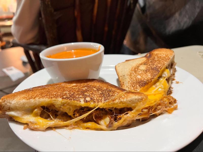 A grilled cheese sandwich with pork inside. In the background is a tomato soup.
