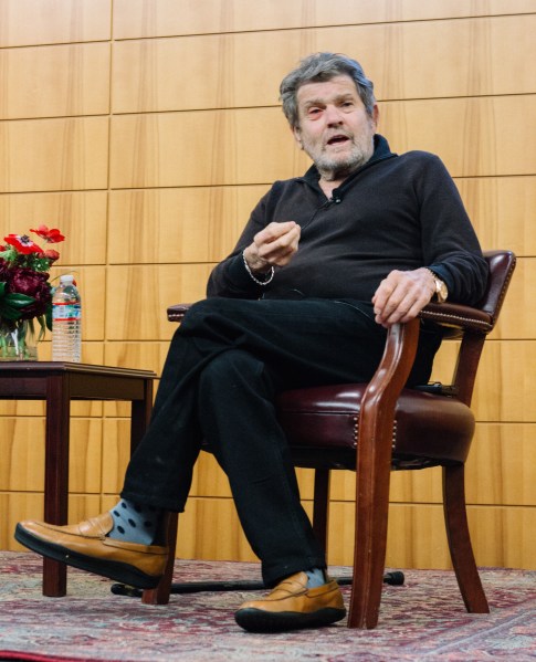 Wenner sits in an all-black outfit with polka dot socks in a wooden chair on stage, in the middle of talking.