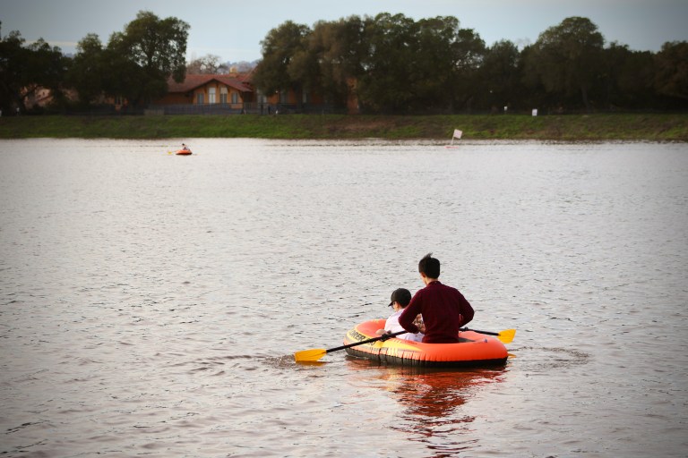 Two people in a bright orange inflatable kayak with yellow paddles sit on the lake. The green banks of the lake in the background.