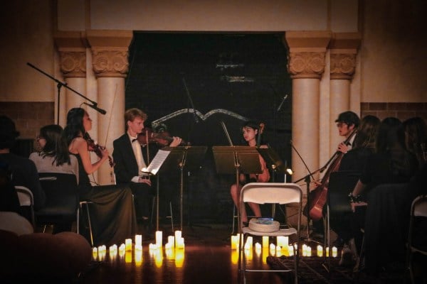a photo of four string players dressed in black formalwear and seated on a dim, candlelit stage, playing their instruments
