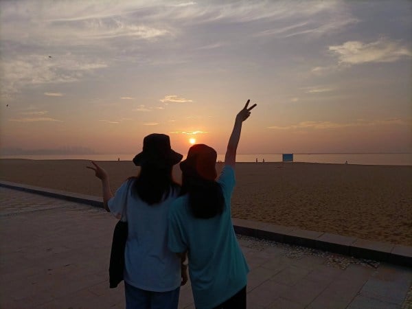 Two sisters watch the sunrise, standing together on a beach. Both have an arm raised in a peace sign.