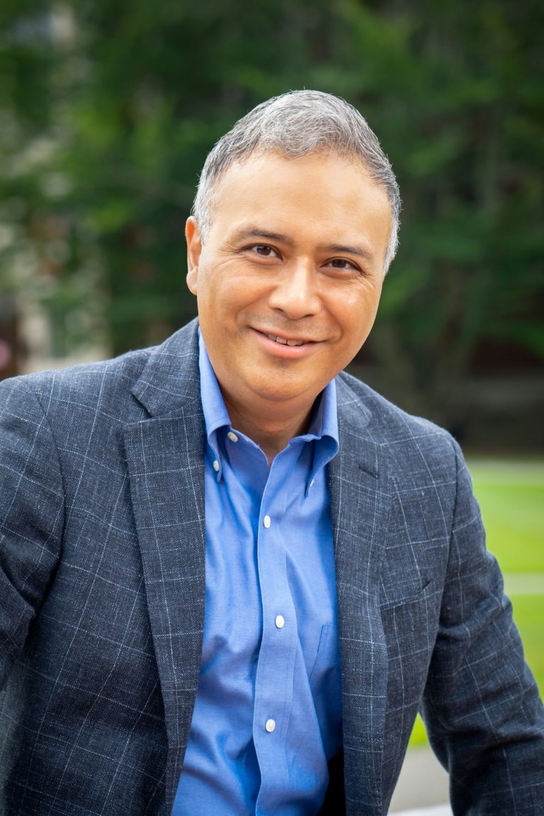 a portrait photograph of a smiling masculine person with tan skin and gray hair; the person is wearing a blue dress shirt and gray jacket