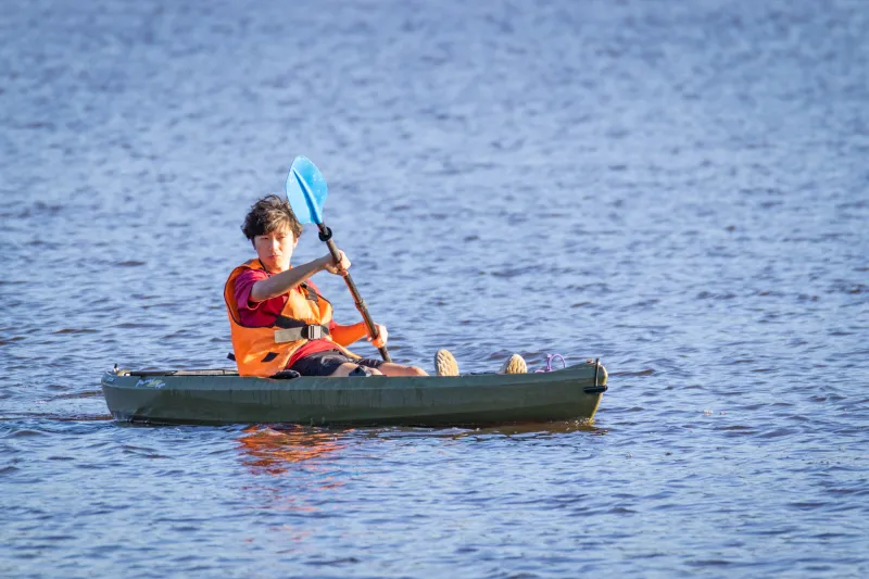 A student wearing an orange life vest holds a paddle in a green kayak.
