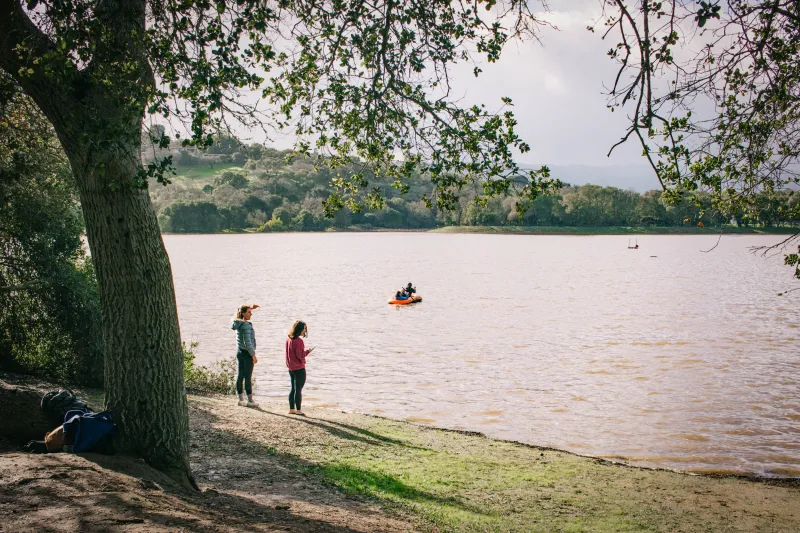 Two friends stand at the edge of Lake Lagunita, as two people paddle in an inflatable orange kayak across the lake in the background.