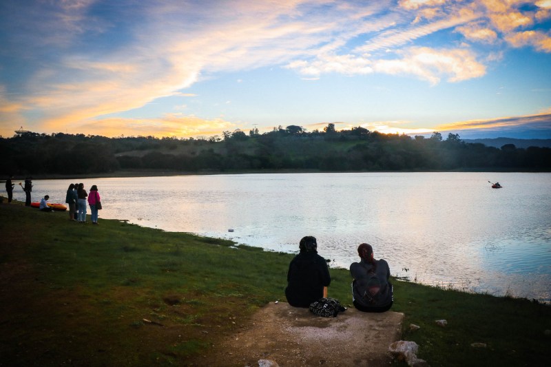 Two students sit with their backs facing the camera looking at the lake at sunset.