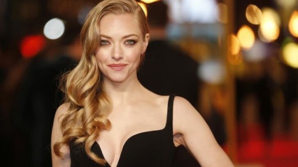 Amanda Seyfried stands in front of a red carpet event in an elegant black dress.